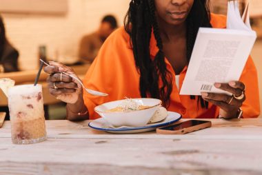 a cropped image of an African-American woman reading a book while eating a dinner